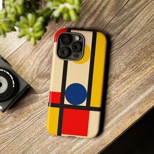 May include: A phone case with a colorful geometric design. The case features a cream background with a red square, a blue circle, and yellow and black vertical stripes. The phone case is on a wooden surface.