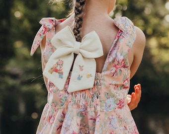 Op de afbeelding: Een klein meisje draagt een roze bloemenjurk met een witte strik die aan de achterkant is gebonden. De strik heeft een geborduurde letter "A" met kleurrijke bloemen en een paar kleine geborduurde bloemen op de strik.