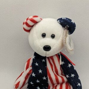 May include: A white plush bear with red and blue stripes and stars, resembling the American flag. The bear has black eyes and a black nose. The bear is wearing a tag that says "Ty Inc."