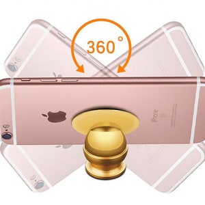 Może przedstawiać: Złoty magnetyczny uchwyt do telefonu z obrotową kulką o 360 stopniach. Uchwyt jest przymocowany do różowego iPhone'a.