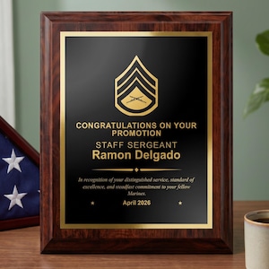 May include: A rectangular wooden plaque with a dark brown finish and a black metal plate. The plate features a gold Staff Sergeant rank insignia, the text CONGRATULATIONS ON YOUR PROMOTION, STAFF SERGEANT Ramon Delgado, and the date April 2026.