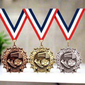 May include: Three soccer medals with red, white, and blue striped ribbons. The medals are bronze, gold, and silver, each featuring a soccer ball, cleat, and net design within a starburst frame. The medals are displayed on a white surface.