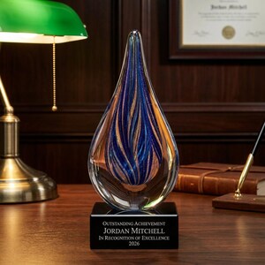 Op de afbeelding: Een druppelvormige glazen trofee met wervelende blauwe en gouden patronen aan de binnenkant, gemonteerd op een zwarte rechthoekige voet. De voet is gegraveerd met de tekst: OUTSTANDING ACHIEVEMENT, JORDAN MITCHELL, IN RECOGNITION OF EXCELLENCE, 2026.