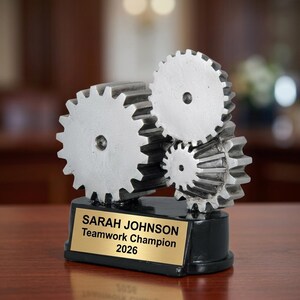 以下が含まれることがあります： 黒い台座に銀色の歯車彫刻。 金色のプレートには「SARAH JOHNSON Teamwork Champion 2026」と書かれています。 歯車は連動しており、チームワークを連想させます。 背景はぼやけています。