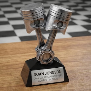 Puede incluir: Un trofeo plateado con dos pistones de motor unidos, montado sobre una base negra. La base tiene una placa plateada con el texto "NOAH JOHNSON OMAHA CARS AND COFFEE 2026 BEST IN SHOW". El trofeo está sobre una superficie de madera.