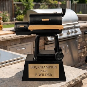 May include: A black BBQ Champion trophy with a miniature grill design. The trophy has a wooden shelf and a gold plaque that reads "BBQ CHAMPION 2026 P. WILDER". The trophy is set on a stone surface.