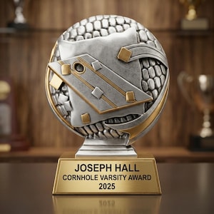 May include: A silver and gold-toned trophy featuring a textured circular emblem with a cornhole board graphic. The base is a rectangular gold plaque with the engraved text: JOSEPH HALL, CORNHOLE VARSITY AWARD, 2025. The design includes metallic accents.
