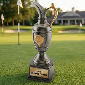 May include: A dark gray golf trophy with a gold-colored handle and golf ball emblem. The trophy sits on a square base with a gold nameplate that reads "MVP Jordan Smith". The trophy is on a green golf course.