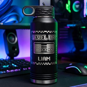 May include: Black water bottle with a black lid and handle. The bottle has a silver design that says "LEVEL UP!" with a controller graphic and the name "LIAM". The bottle is designed for gamers.