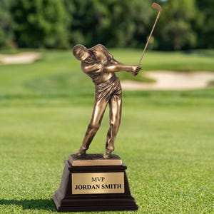 May include: A bronze-colored golf trophy featuring a golfer in mid-swing. The trophy is mounted on a dark brown base with a gold-colored plate that reads "MVP JORDAN SMITH". The background is a green golf course.