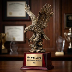 May include: A bronze eagle statue with outstretched wings, perched on a rocky base, atop a wooden trophy base. The base has a gold plaque that reads "MICHAEL DAVIS OUTSTANDING PATRIOTIC 2025". The background includes framed certificates and trophies.