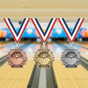 May include: Three bowling medals with red, white, and blue striped ribbons. The medals are bronze, gold, and silver, each featuring a bowling pin and ball design surrounded by stars. The background shows a bowling alley.
