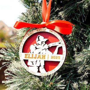 May include: Red and wood-tone Christmas ornament with a shooting sports theme. The ornament features a silhouette of a person aiming a rifle, the name "ELIJAH", and the year "2025". The text "NO-TARGET, NO-BIRD", "BROKEN TARGET", "ALL-AROUND FIVE STAND", and "TRAP MACHINE" are also included.