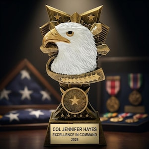 Puede incluir: Un trofeo dorado con una cabeza de águila blanca, pico y plumas detalladas. El trofeo tiene un emblema en forma de estrella y el texto "COL JENNIFER HAYES EXCELLENCE IN COMMAND 2026" en una placa dorada. El fondo incluye una bandera americana plegada y medallas.