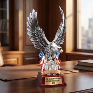 May include: A silver-colored eagle statue with outstretched wings, perched atop a base with an American flag design. The base has a gold plaque that reads "SSG CARLOS MARTINEZ MISSION EXCELLENCE 2025". The statue is on a wooden desk.