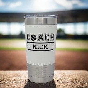 May include: White stainless steel tumbler with a white leatherette wrap. The wrap has black text that reads "COACH NICK" with a baseball graphic.