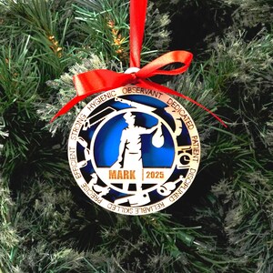 May include: A wooden Christmas ornament with a red ribbon. The ornament features a silhouette of a person holding a bag, with the name "MARK" and the year "2025" below. The outer ring has words like "HYGIENIC" and "EFFICIENT".