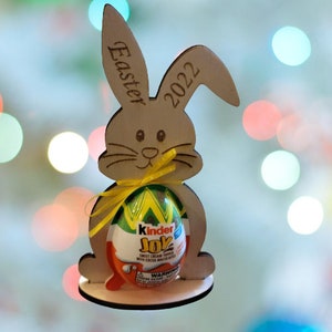 Puede incluir: Corte de conejo de Pascua de madera que sostiene un huevo de chocolate Kinder Joy. El conejo está decorado con las palabras "Easter 2022".