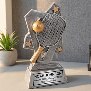 May include: Silver and gold pickleball trophy with a paddle and ball design. The trophy base has a plaque that reads "NOAH JOHNSON OMAHA PICKLEBALL LEAGUE 2026 CHAMPION". Includes gold stars.
