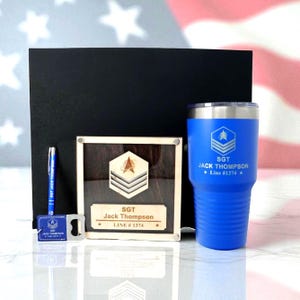 May include: A gift set featuring a blue tumbler, a wooden plaque, a pen, and a bottle opener. The tumbler and pen have the text "SGT JACK THOMPSON Line #1374". The wooden plaque has a military insignia and the same text. The set is presented in a black box.