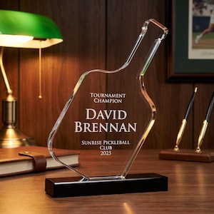 Könnte beinhalten: Eine transparente Acryl-Trophäe in Flaschenform mit schwarzem Sockel. Die Trophäe ist mit den Worten "Tournament Champion", "David Brennan", "Sunrise Pickleball Club" und dem Jahr "2025" graviert.