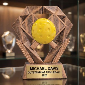 Può includere: Un trofeo di pickleball color bronzo con una pallina gialla e racchette incrociate. Il trofeo ha una cornice a forma di diamante e una base rettangolare con la scritta "MICHAEL DAVIS OUTSTANDING PICKLEBALL 2025".
