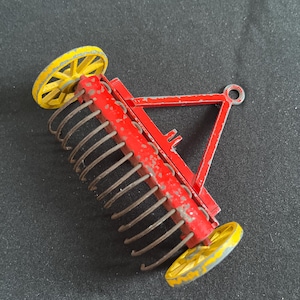 Op de afbeelding: Een vintage speelgoed boerenhark, met een rood metalen frame, gebogen tanden en twee gele wielen. Het speelgoed heeft een trek-ontwerp en staat tegen een donkere achtergrond. De hark is ongeveer 15 cm lang.