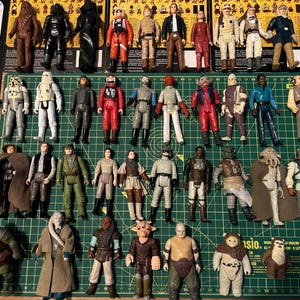 May include: A selection of Star Wars action figures, including Darth Vader, Chewbacca, and other characters from the series. The figures are displayed on a green cutting mat, with some still in their original packaging.
