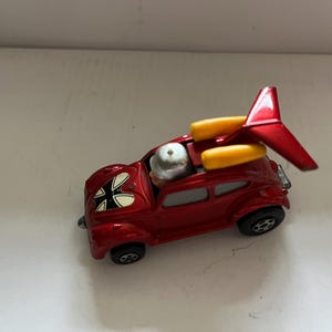 May include: A red toy car with a black and white design on the bonnet. The car has a yellow and red surfboard attached to the roof. The car has a small figure of a person in the driver's seat.