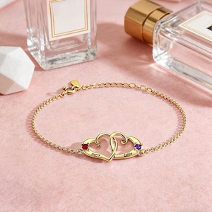 May include: A gold-colored bracelet with two intertwined heart-shaped pendants. Each heart has a gemstone, one red and one purple. The bracelet has the names "Herry" and "Callie" engraved on the hearts. The background includes perfume bottles.