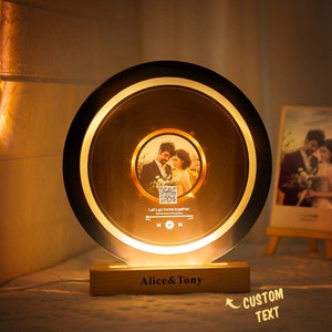 May include: A round, illuminated photo frame with a couple's photo and a QR code. The frame has a wooden base with the text "Alice & Tony" engraved on it. The frame is lit with warm white light.