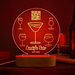 Op de afbeelding: Een rood LED-licht met een houten basis heeft een transparant acryl paneel met een gele omtrek van een martini-glas en andere glazen, een QR-code en de tekst "Andy's Bar EST 2023".