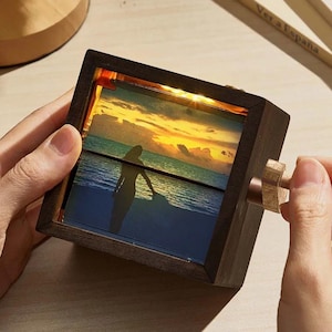 May include: A wooden picture frame with a built-in light, featuring a photo of a sunset over the ocean with a silhouette of a person standing on the beach. The frame has a crank on the side to rotate the photo.