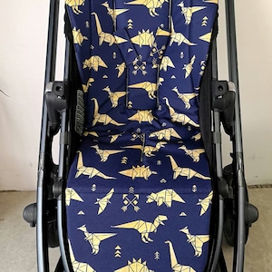 May include: A black stroller with a navy blue and gold dinosaur patterned seat cover. The seat cover features origami-style dinosaurs in a geometric pattern.