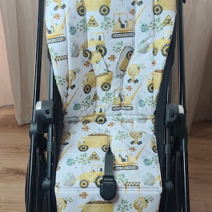 Könnte beinhalten: Ein weißer Kinderwagen-Sitzbezug mit einem gelben und grünen Druck von Baufahrzeugen. Der Druck zeigt verschiedene Baufahrzeuge, darunter Kipplaster, Bagger und Bulldozer. Der Text "Duck the Paint" wird auf dem Druck wiederholt.