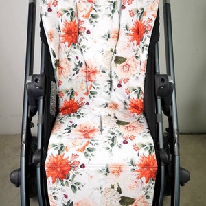 May include: A pushchair seat with a floral design. The seat has a white base with large orange and pink flowers, green leaves, and small purple accents. The pushchair frame is black and grey with brown leather detailing.