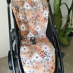 Könnte beinhalten: Ein beigefarbener Kinderwagen-Sitzbezug mit einem Blumenmuster mit weißen, orangefarbenen und blauen Blumen. Der Bezug ist aus einem weichen, atmungsaktiven Stoff gefertigt und passt auf die meisten Standard-Kinderwagen-Sitze.
