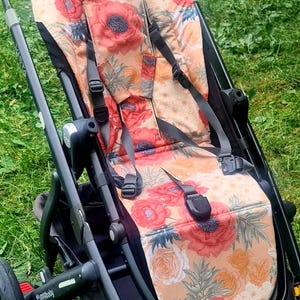 Könnte beinhalten: Schwarzer Kinderwagen mit einem Sitz mit Blumenmuster mit großen roten und orangen Blumen auf beigem Hintergrund. Der Kinderwagen hat einen braunen Griff und schwarze Räder, vor einem grünen Gras Hintergrund.