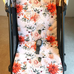 Può includere: Una fodera per seggiolino passeggino bianca con un motivo floreale con fiori arancioni, rosa e verdi. La fodera ha uno sfondo bianco con un motivo floreale.