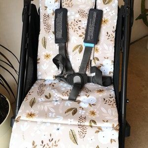 Könnte beinhalten: Ein Kinderwagensitz mit beigem Blumenmuster. Das Muster zeigt weiße und hellblaue Blumen sowie braune und goldene Blätter. Der Kinderwagen hat einen schwarzen Rahmen und einen grauen Gurt.