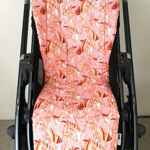 May include: A pink and white stroller seat cover featuring a pattern of pink cockatoos. The cover is made from a soft, comfortable fabric and is designed to fit most standard stroller seats.