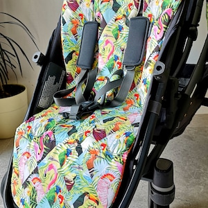 Könnte beinhalten: Ein schwarzer Kinderwagen mit einer bunten Sitzbezug mit tropischem Vogelmuster. Der Bezug zeigt Tukane, Papageien und rosa Flamingos auf einem grün-blauen Hintergrund.