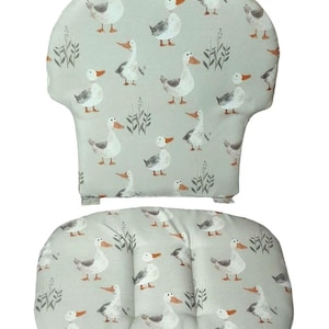 Könnte beinhalten: Ein Set aus zwei Stuhlkissen mit hellgrünem Hintergrund und einem Muster aus grauen und weißen Enten. Die Kissen sind oval geformt und haben ein gestepptes Design.