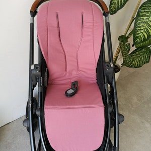 Könnte beinhalten: Ein rosa Kinderwagen-Sitz mit schwarzem Gestell und braunen Lederakzenten. Der Sitz hat eine gepolsterte Rückenlehne und einen Fünf-Punkt-Gurt.