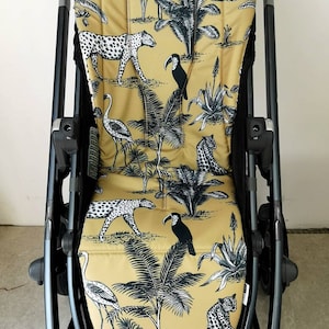 May include: A stroller seat cover with a jungle print featuring toucans, leopards, flamingos, and palm trees on a mustard yellow background.