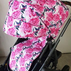 Könnte beinhalten: Ein schwarzer Kinderwagen mit einem rosafarbenen floralen Verdeck und Sitzbezug. Das Verdeck hat einen weißen Hintergrund mit rosafarbenen Pfingstrosen und grünen Blättern. Der Sitzbezug hat das gleiche Muster.