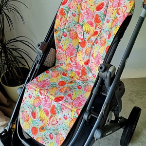Könnte beinhalten: Ein schwarzer Kinderwagen mit einer bunten Sitzbezug mit Fruchtmuster. Der Bezug zeigt Ananas, Bananen, Erdbeeren, Wassermelonen und Zitrusfrüchte auf hellblauem Hintergrund.