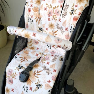 Könnte beinhalten: Ein weißer Kinderwagen-Sitzbezug mit einem Blumenmuster in Rosa-, Braun- und Beigetönen. Der Bezug zeigt eine Vielzahl von Blättern und Blumen, darunter kleine rosa Blüten, große beige Blüten und braune Blätter.