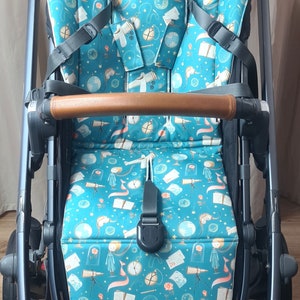 May include: A black stroller with a teal and white patterned seat cover featuring a whimsical design of a little prince, a plane, and a globe. The stroller has a brown leather handle and a black mesh basket.