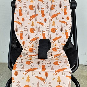 Könnte beinhalten: Ein beiger Bezug für den Sitz eines Kinderwagens mit einem sich wiederholenden Muster aus orangefarbenen Füchsen und braunen Bäumen. Der Bezug ist aus einem weichen, komfortablen Stoff gefertigt und soll den Sitz des Kinderwagens vor Schmutz und Verschüttungen schützen.